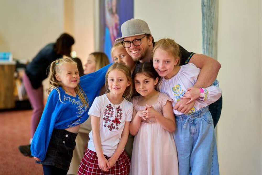 A group of five young girls smiling and posing for a photo with an adult man wearing glasses and a grey cap. The group appears cheerful and friendly, gathered in a bright indoor space with others in the background.