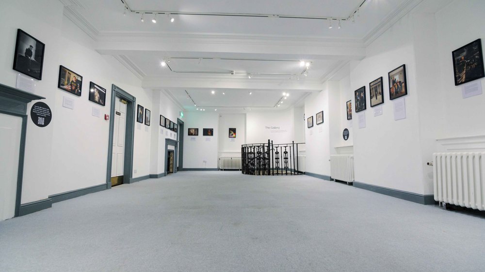 A bright gallery space with framed photographs lining both walls, leading towards a circular metal railing at the back, which surrounds a staircase descending to a lower level. 