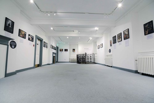 A bright gallery space with framed photographs lining both walls, leading towards a circular metal railing at the back, which surrounds a staircase descending to a lower level.