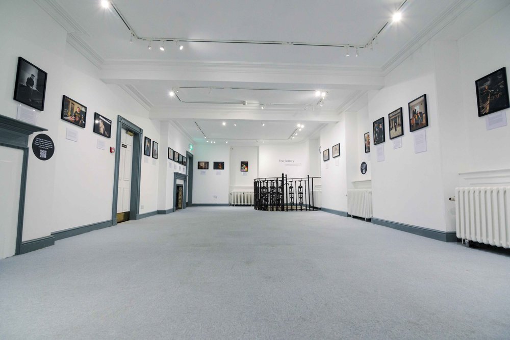 A bright gallery space with framed photographs lining both walls, leading towards a circular metal railing at the back, which surrounds a staircase descending to a lower level. 