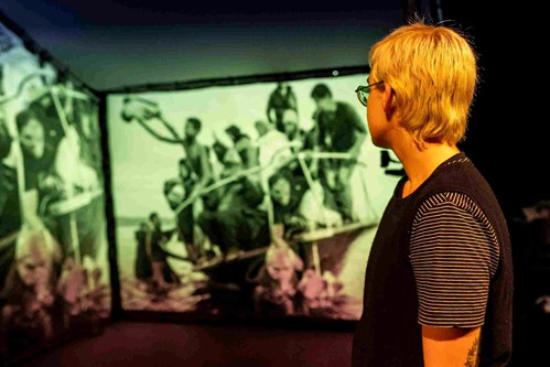 A young person stands looking at a projection behind them. The projection is an image of a small boat with too many people in it. 