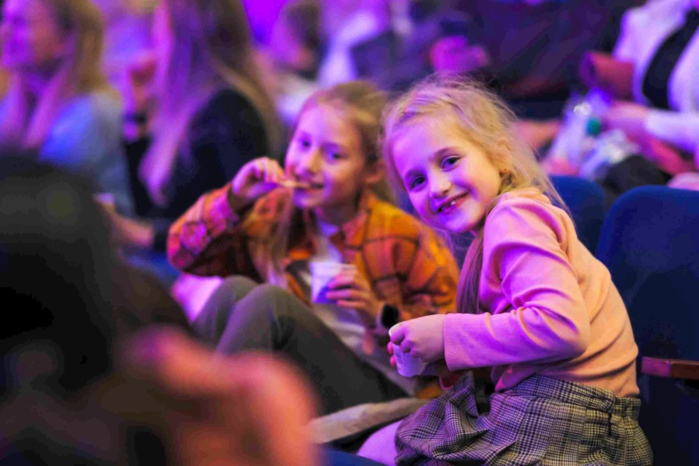 Two young girls sitting in theatre seats, enjoying snacks while smiling and looking towards the camera. 