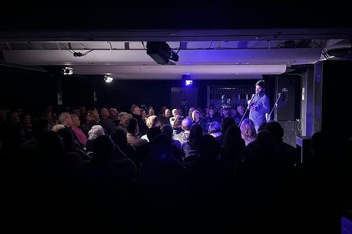 A comedian stands on stage performing to a packed audience in a dimly lit indoor theatre space. He holds a microphone and is mid-speech, with audience members seated closely together and visibly engaged.
