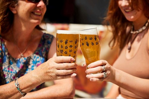 Two women each hold a pint glass and are cheers-ing, with the pint glasses touching. 