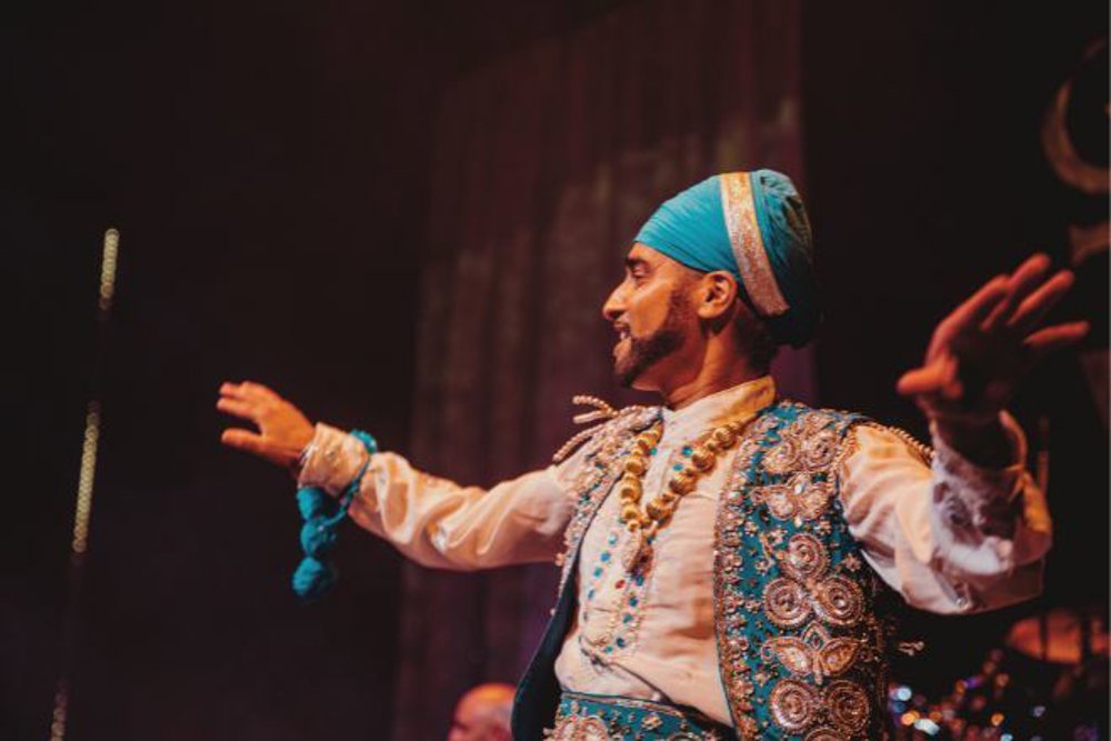 A male performer dressed in traditional Punjabi attire, featuring a bright blue turban and an intricately decorated blue and white vest. He is mid-dance, with arms outstretched and a joyful expression, illuminated by warm stage lighting
