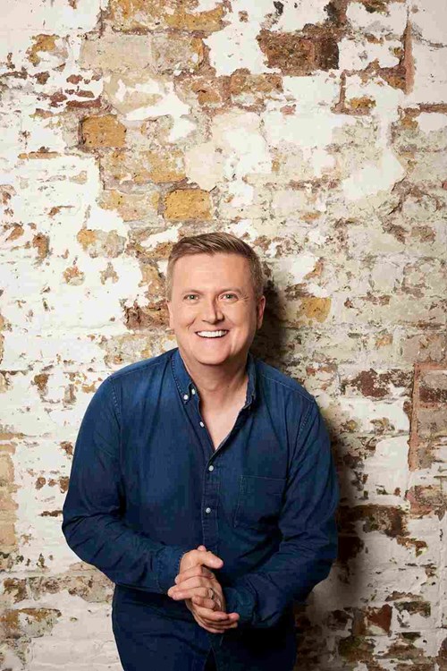 Aled Jones leans against the wall. He is wearing a blue, denim shirt and smiles widely at the camera. 