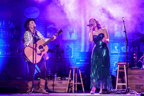 Two female performers sing on stage in dramatic lighting—one plays acoustic guitar and wears a snakeskin jacket and black hat, while the other, in a shimmering teal dress, sings into a microphone.