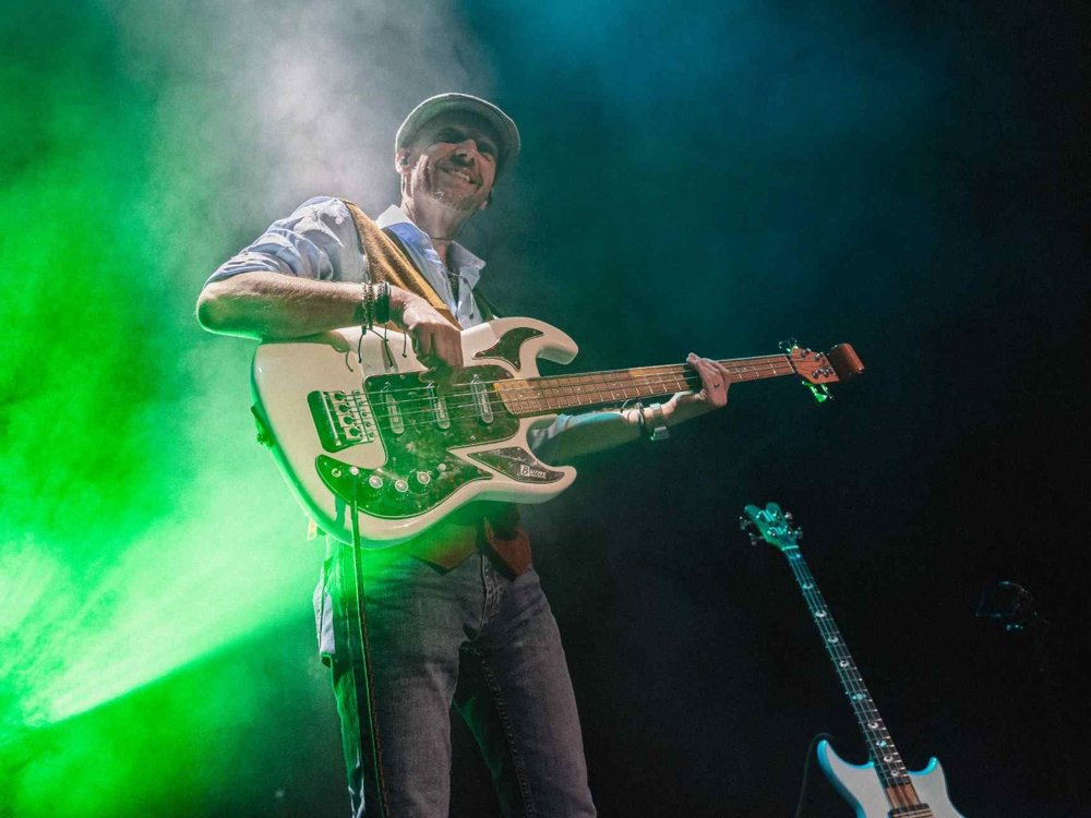 A musician smiling while playing a white electric bass guitar, standing in bright green and blue stage lighting with atmospheric smoke in the background and another guitar visible behind them.