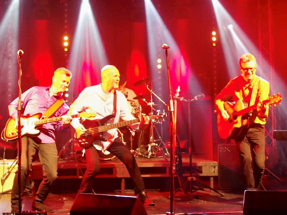 Four-piece band performing energetically on stage under bright red and white lights, playing electric guitars and drums during a live 1960s tribute show