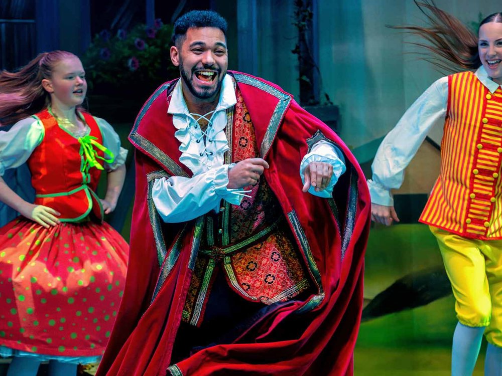 A smiling male lead runs forward mid-dance, wearing a red cape and patterned waistcoat, joined by two younger performers in bright storybook costumes behind him.