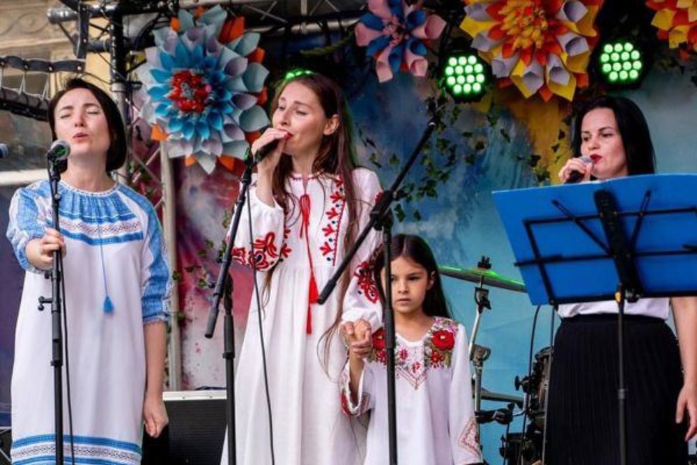 Four female performers, including a young girl, stand on a decorated stage singing into microphones. They are wearing traditional embroidered clothing, and vibrant oversized paper flowers hang behind them, lit by green stage lights.