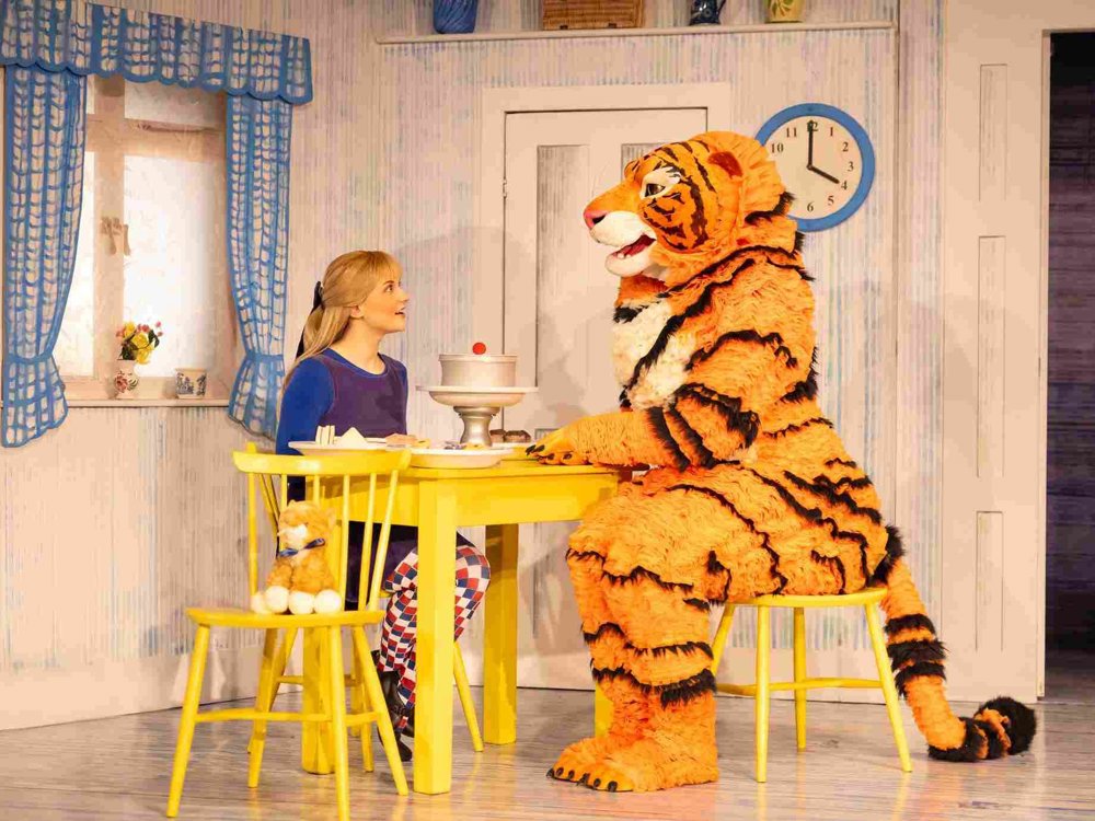 A young woman sits at a yellow kitchen table facing a large friendly tiger character in costume, with a cake on a stand between them on a pastel blue and white stage set.