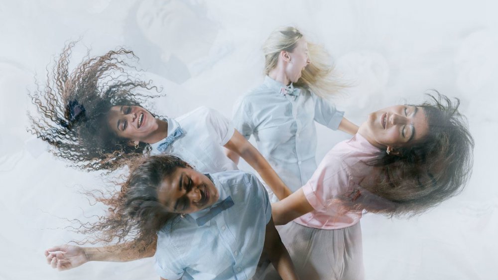 An image of four dancers in pastel shirts and bow ties move expressively with flowing hair and joyful expressions against a soft, misty background.
