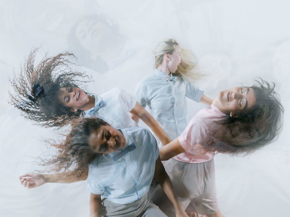 An image of four dancers in pastel shirts and bow ties move expressively with flowing hair and joyful expressions against a soft, misty background.