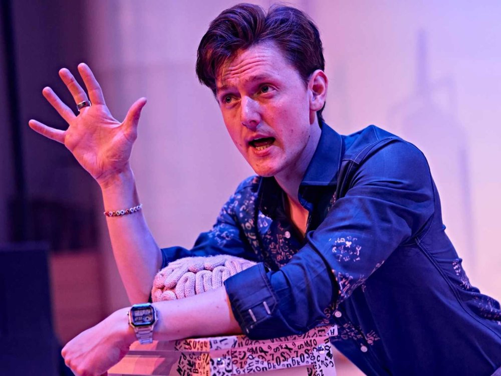 A performer in a blue patterned shirt sits on stage under pink and purple lighting, gesturing expressively with one hand while speaking. Leaning on a stack of props, they appear to be engaged in a storytelling or theatrical performance. The background is softly lit, focusing attention on the performer and their animated expression.