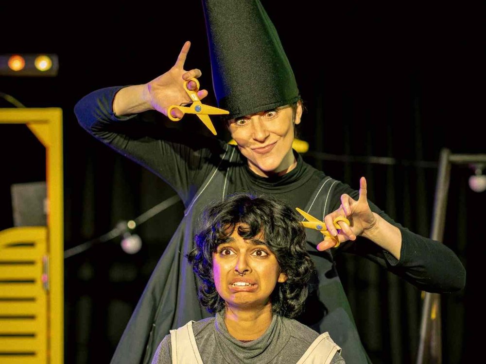 A performer dressed as a witch stands behind Rapunzel, holding oversized yellow scissors beside her long hair. Rapunzel looks alarmed as the pair strike a comic pose on stage.