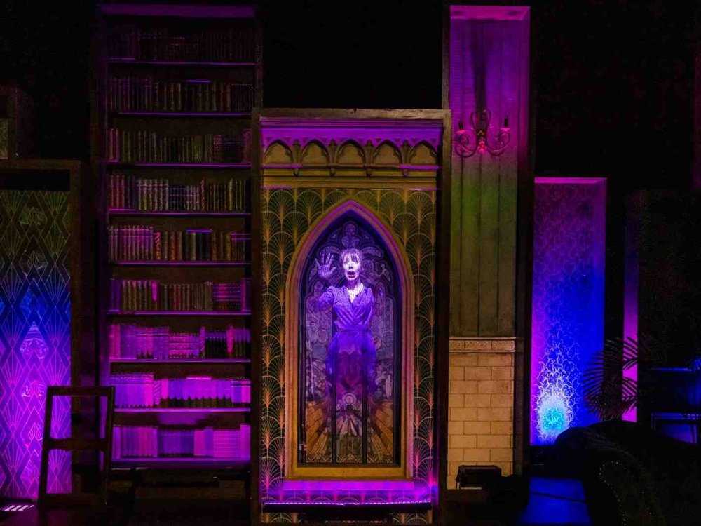 A haunting stage image with purple and blue lighting showing a performer’s ghostly projection framed by gothic-style bookcases and stained glass.