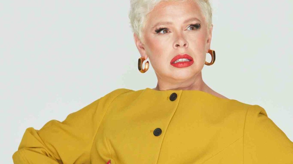 A person with short platinum blonde hair wearing a mustard yellow buttoned jacket, gold hoop earrings, and bold red lipstick, striking a dramatic pose with hands on their waist against a plain light background.
