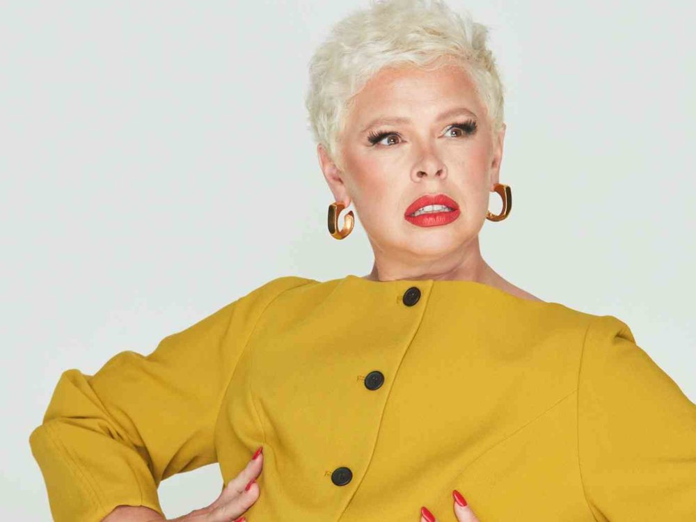 A person with short platinum blonde hair wearing a mustard yellow buttoned jacket, gold hoop earrings, and bold red lipstick, striking a dramatic pose with hands on their waist against a plain light background.