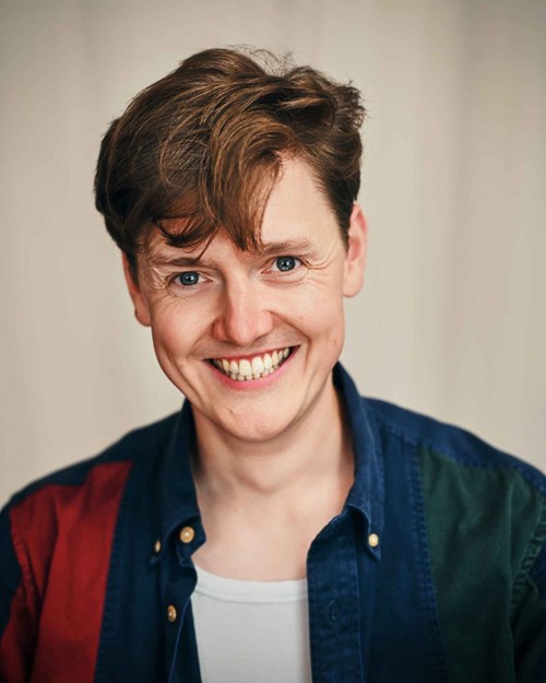 A smiling person with short, light brown hair, wearing a blue and red button-up shirt over a white top, looking directly at the camera against a neutral background.