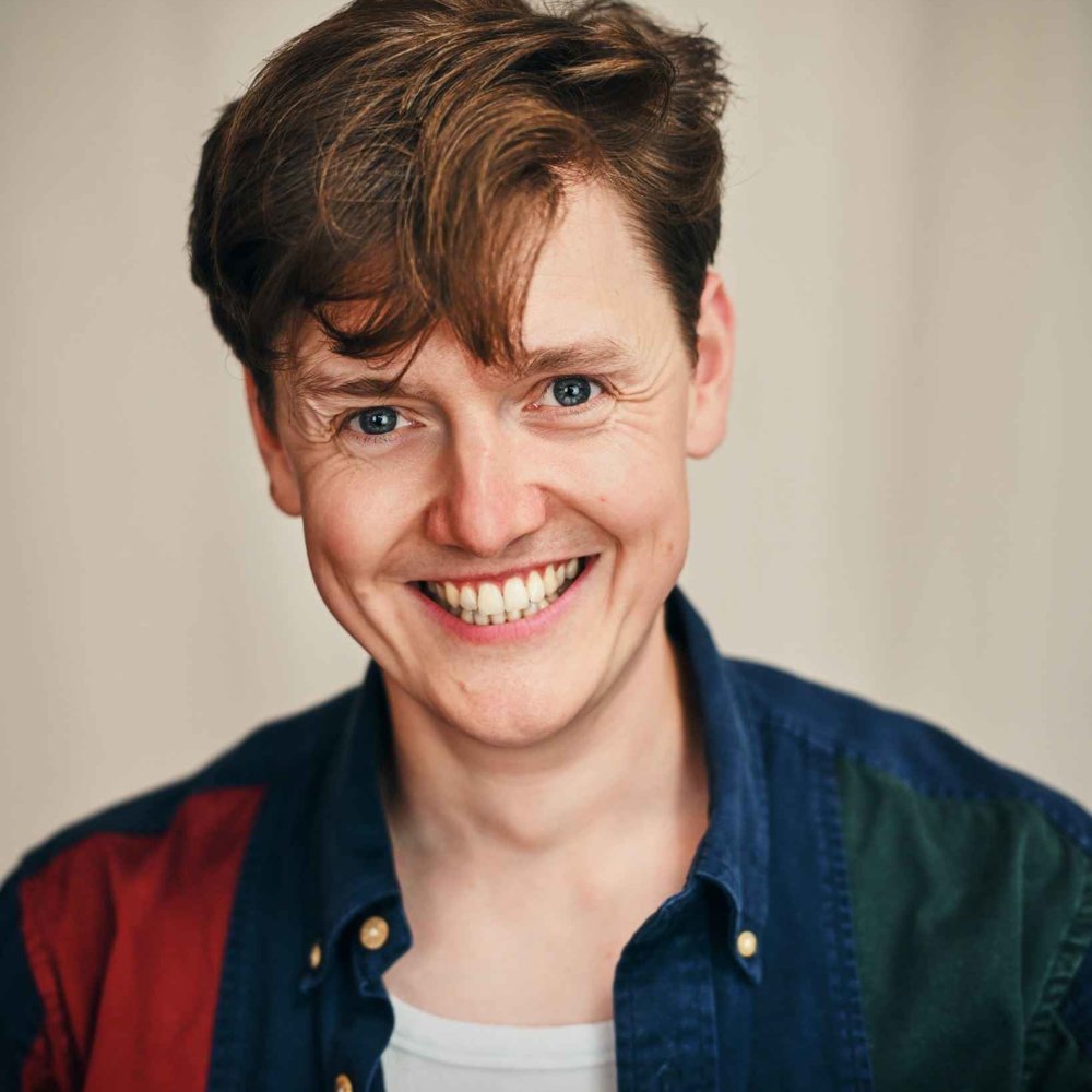 A smiling person with short, light brown hair, wearing a blue and red button-up shirt over a white top, looking directly at the camera against a neutral background.