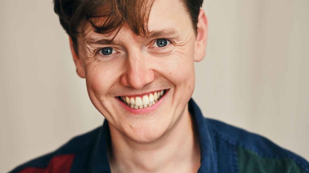 A smiling person with short, light brown hair, wearing a blue and red button-up shirt over a white top, looking directly at the camera against a neutral background.