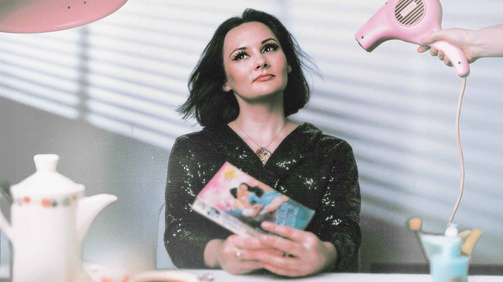 A woman with dark shoulder-length hair wearing a black sequined top looks upward while holding a book. A hand holds a pink hairdryer beside her, with a teapot and other objects blurred in the foreground.