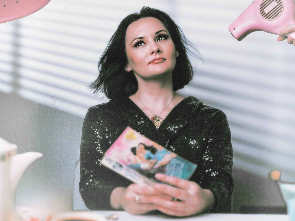 A woman with dark shoulder-length hair wearing a black sequined top looks upward while holding a book. A hand holds a pink hairdryer beside her, with a teapot and other objects blurred in the foreground.