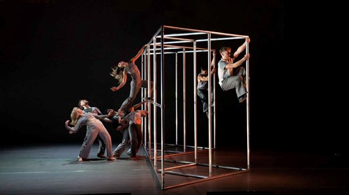 A visually compelling scene from Hidden by Motionhouse, featuring performers climbing and balancing on a large metal cube structure. Other dancers on the stage interact with each other in expressive poses, adding to the energy of the moment. Credit: Dan Tucker.