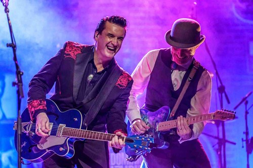 Two smiling male guitarists play electric guitars side by side. One wears a stylish black jacket with red embroidery, the other a vest and bowler hat. Both are lit by vibrant purple and blue stage lights.