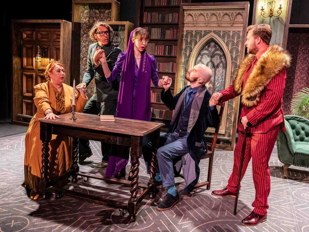 Five actors gathered around a wooden table mid-scene, reacting dramatically; the set features vintage furniture, patterned wallpaper, and a stained-glass window backdrop.