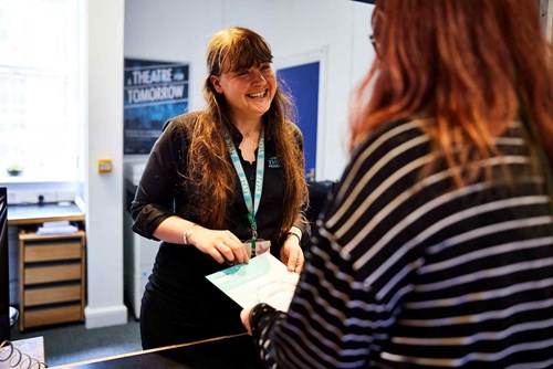 A woman with long brown hair wearing a black shirt and a light blue lanyard smiles at a customer. 