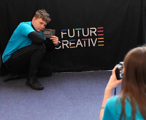 A young man posing with a Future Creative postcard in front of a Future Creative branded black backdrop while a young woman takes his picture
