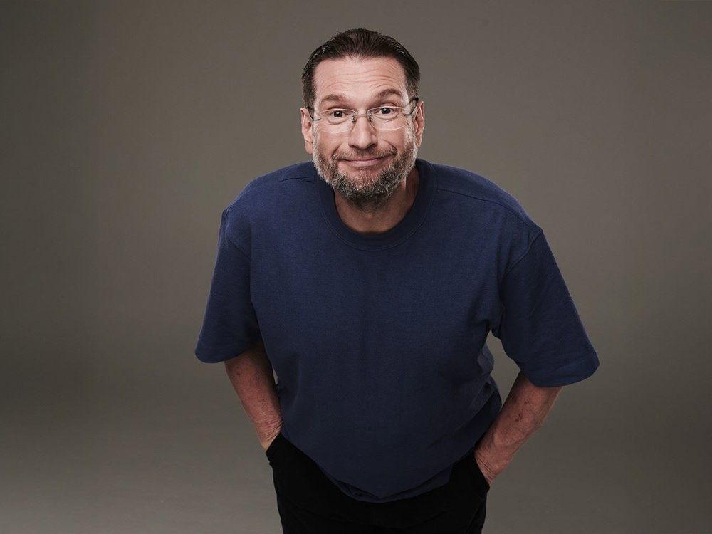 Comedian Gary Delaney stands against a bright blue background, smiling confidently in a dark jacket and shirt, promoting his upcoming tour.