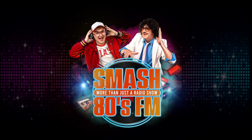 Two presenters dressed in retro 1980s style stand in front of a neon graphic. One wears a red jacket and white cap, the other a curly wig, sunglasses, and a white suit jacket. Between them is a glowing circular logo that reads “SMASH 80’s FM – More Than Just a Radio Show,” surrounded by cassette tapes and colourful lights.