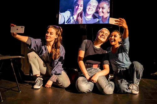 Sitting down, three young people hold their phones out in front of them. Behind them there is a screen, showing their faces as they make the video call. Two young people are sitting close together and smiling. Another young person sits away from the pair looking more serious. 