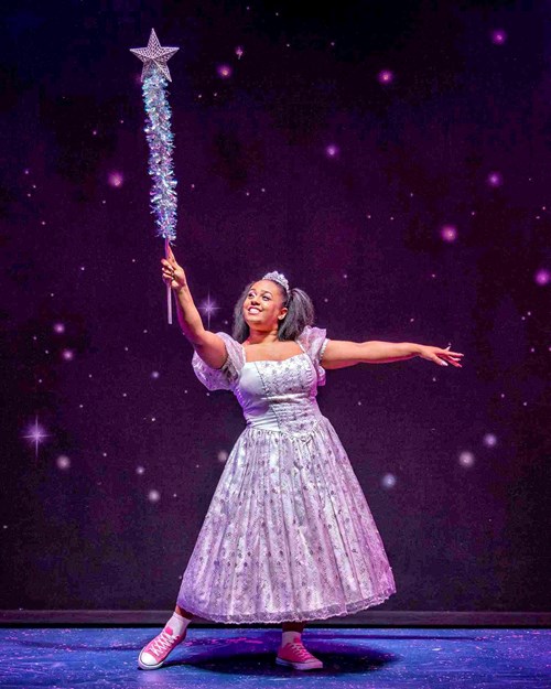 The Fairy Godmother poses with her magical wand. She is wearing a tiara with a long dress that's patterned with sequins.