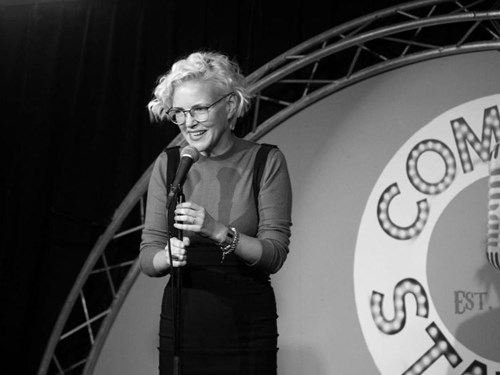 Comedian Ruth Cockburn stands on stage holding a microphone. She is smiling. Ruth has short, blond, wavy hair and is wearing glasses. 