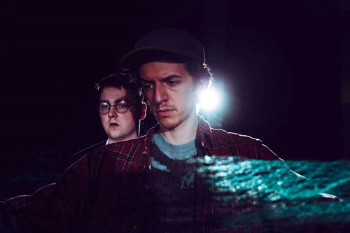 Two performers in a dark space, both looking away from the camera with faces of concern. 