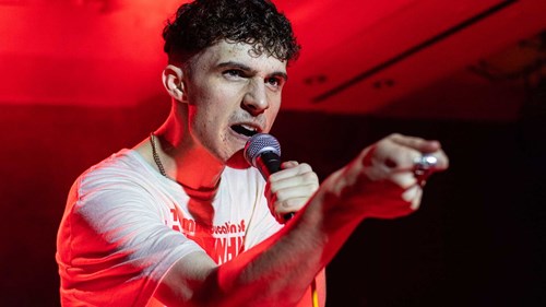 A young man is performing on stage, pointing forward with a fierce expression while holding a microphone. He is wearing a white T-shirt and is illuminated by red stage lighting.