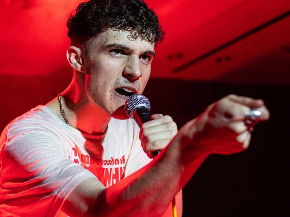 A young man is performing on stage, pointing forward with a fierce expression while holding a microphone. He is wearing a white T-shirt and is illuminated by red stage lighting.