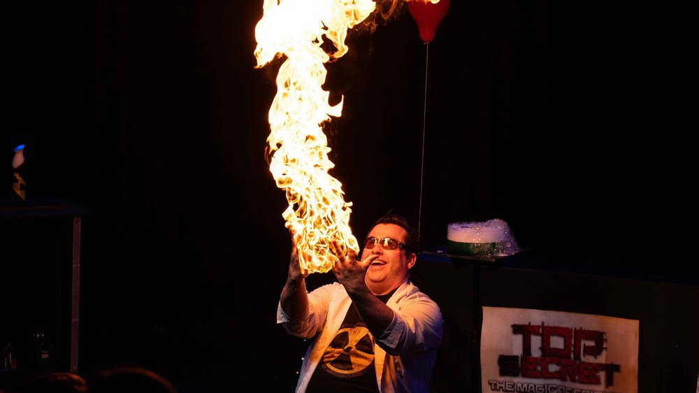 A person in a lab coat creates a tall column of fire during a stage science demonstration.