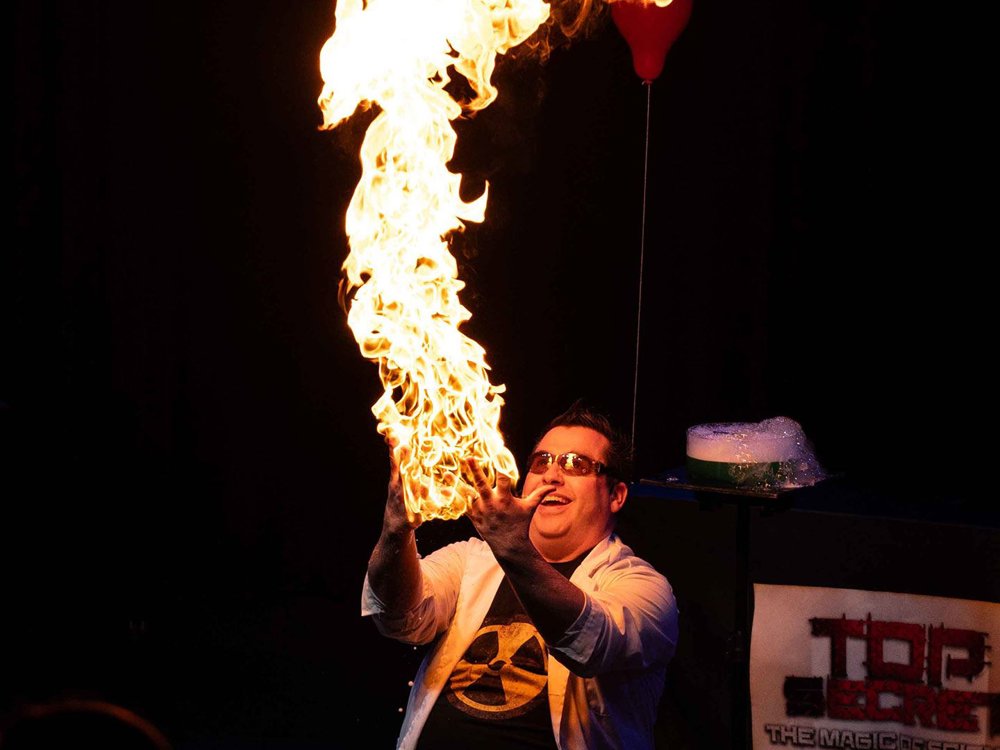 A person in a lab coat creates a tall column of fire during a stage science demonstration.