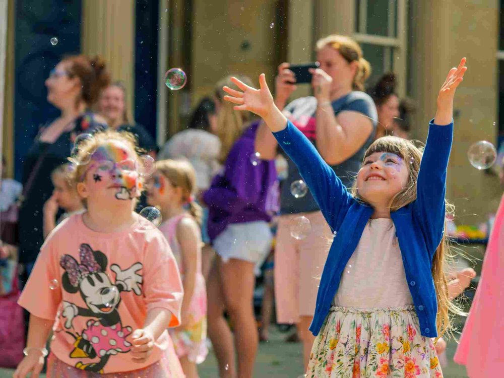 A young girl in a floral skirt and blue cardigan joyfully reaches for bubbles in the air, her face painted with a colourful design. Another child in a pink Minnie Mouse dress watches, surrounded by festival-goers.