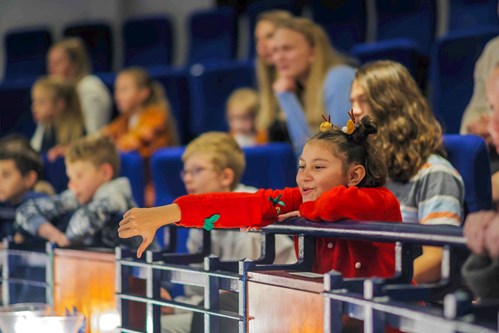 A young girl wearing a festive red jumper with reindeer antlers in her hair leans over a theatre balcony, smiling and giving a playful thumbs-down during a performance, surrounded by other engaged children and adults.