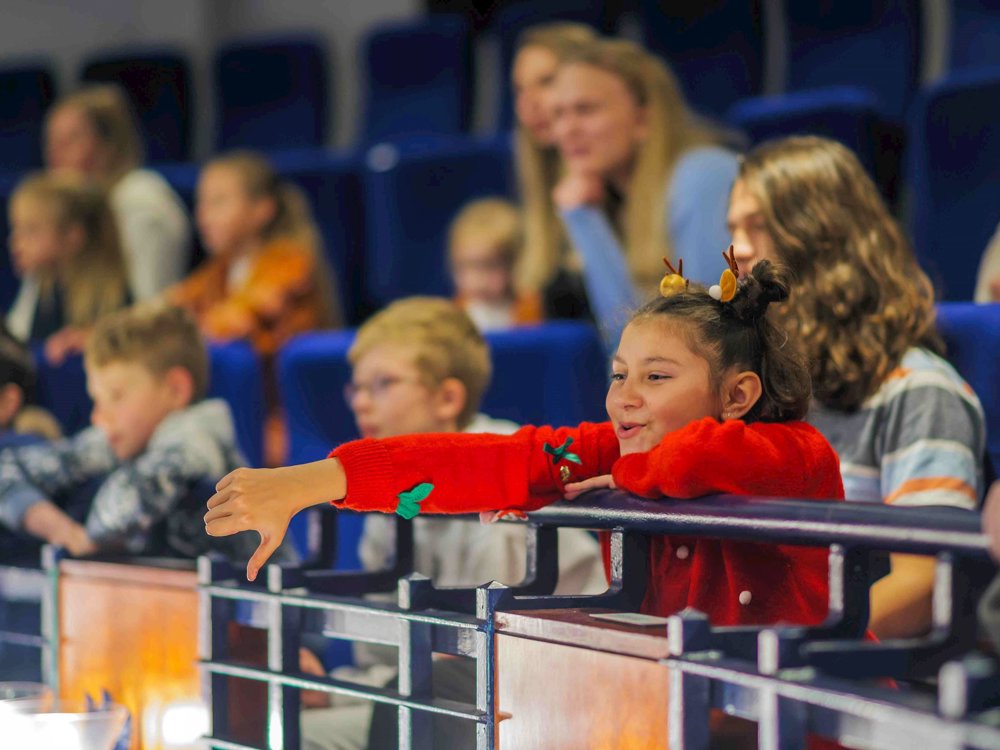 A young girl wearing a festive red jumper with reindeer antlers in her hair leans over a theatre balcony, smiling and giving a playful thumbs-down during a performance, surrounded by other engaged children and adults.