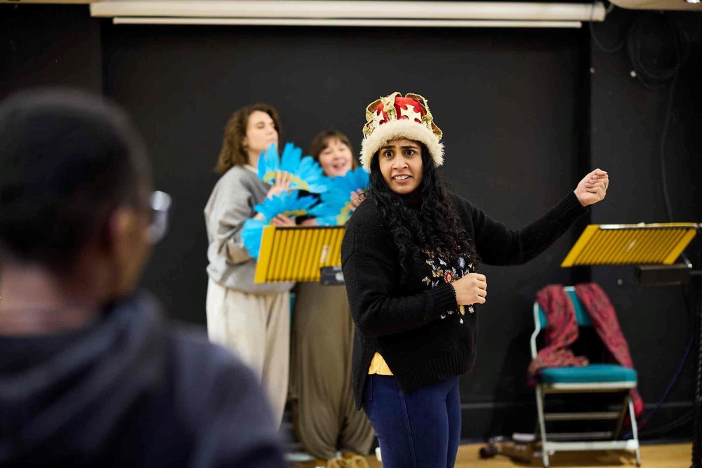 A performer wearing a crown and a black jumper with embroidered details takes centre stage during a rehearsal, gesturing expressively. In the background, two performers hold blue feather fans, standing behind music stands, as another person watches from the foreground.