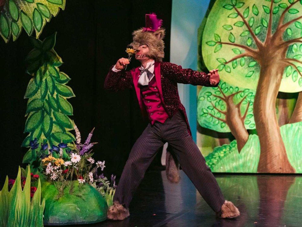 A performer dressed as the Wolf in a red velvet jacket and top hat strikes a dramatic pose on stage, holding a yellow flower in front of a forest backdrop.