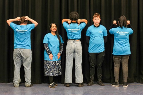 5 young adults in a line, half facing away from the camera all wearing the same blue Future Creative T-shirt, that says Volunteer on the reverse