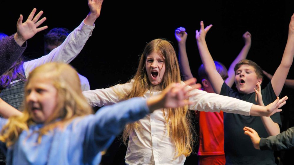 A group of children perform energetically on stage with their arms raised during a theatre workshop or performance. In the centre, a girl with long hair and a white shirt sings or shouts expressively while other children around her strike dramatic poses under colourful stage lighting against a dark background.